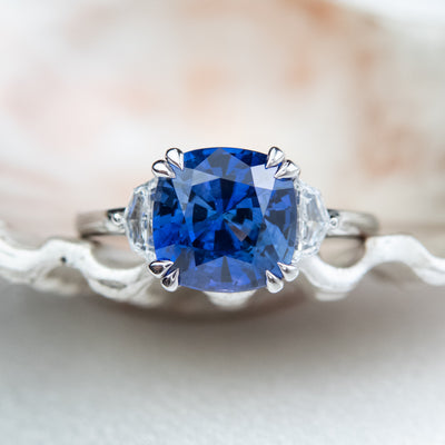 A large cushion shaped blue sapphire ring flanked by white cadillac cut diamonds set in a classic platinum setting against a light, softly blurred background. 