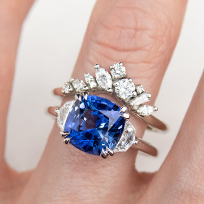 A delicate white gold ring featuring a cluster of sparkling diamonds in various shapes, elegantly stacked with a large blue sapphire cushion cut engagement ring.