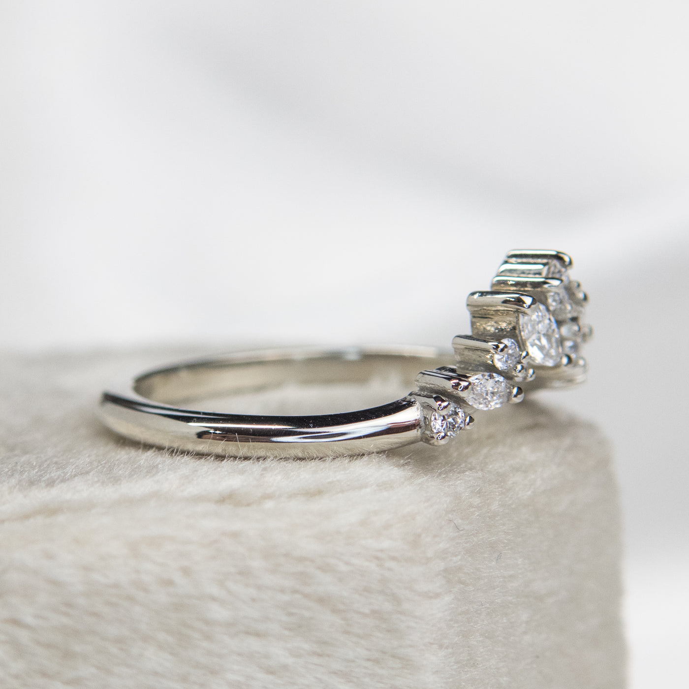 A delicate white gold ring featuring a cluster of sparkling diamonds in various shapes, elegantly set on a soft, light-colored surface shown from the side.