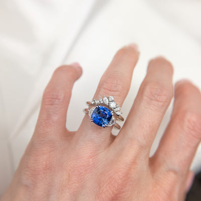 A delicate white gold ring featuring a cluster of sparkling diamonds in various shapes, elegantly stacked on a finger with a large blue sapphire cushion cut engagement ring.