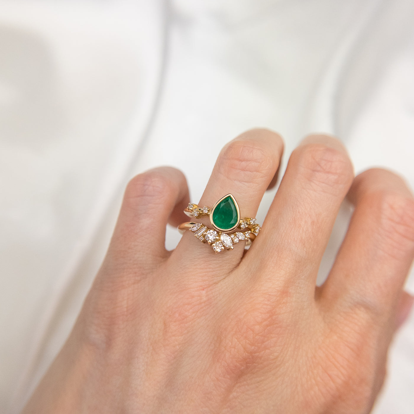 A delicate 14kt gold ring featuring a cluster of sparkling diamonds in various shapes, elegantly stacked on a finger with a pear shaped emerald engagement ring in gold with diamond accents on the band.