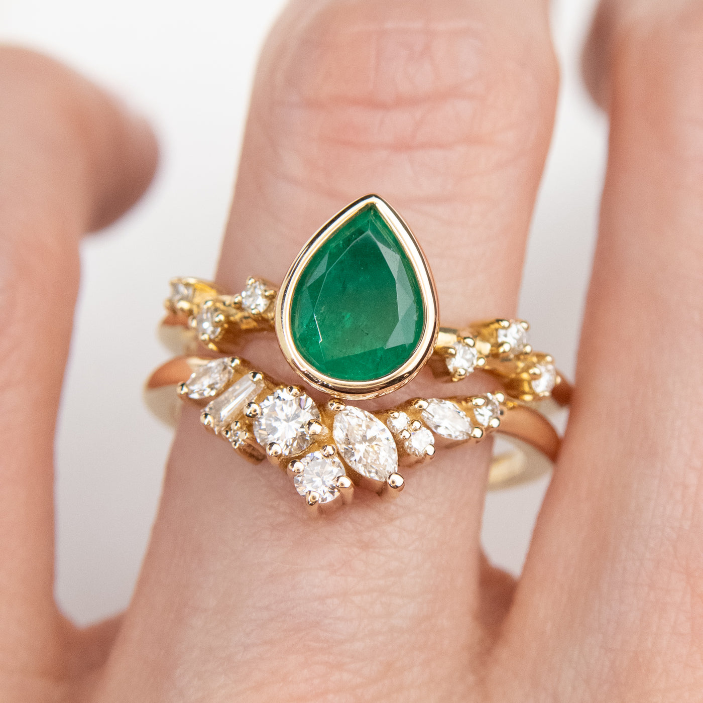 A delicate 14kt gold ring featuring a cluster of sparkling diamonds in various shapes, elegantly stacked on a finger with a pear shaped emerald engagement ring in gold with diamond accents on the band.