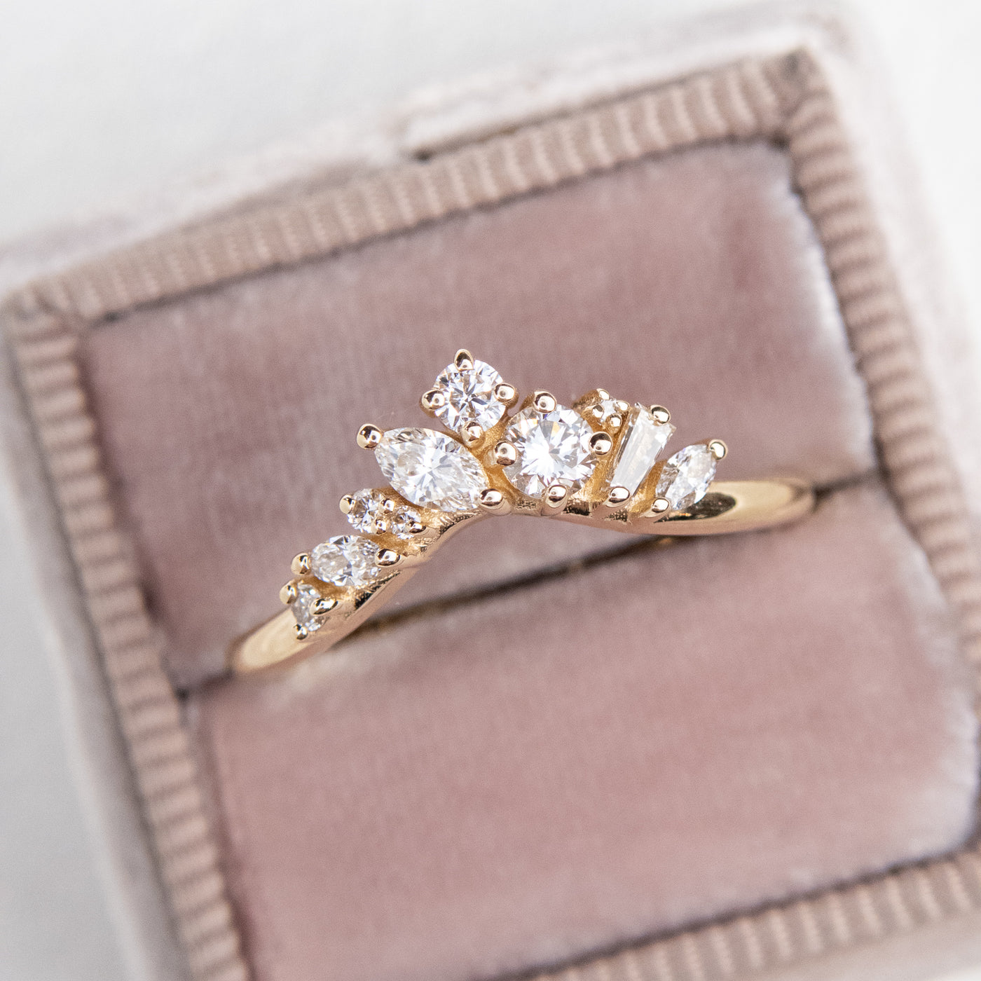 A delicate 14kt gold ring featuring a cluster of sparkling diamonds in various shapes, elegantly resting in a light pink  velvet ring box shown from above.