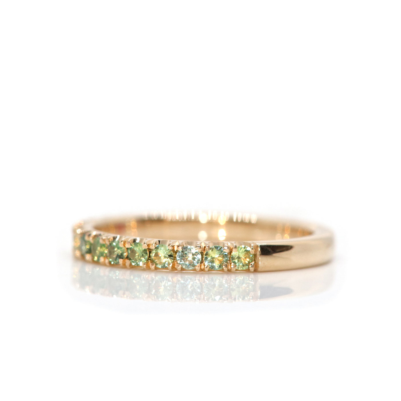 A delicate 14kt gold ring adorned with  brilliant cut parti-green sapphires in a variety of tones, reflecting beautifully against a white background, shown from the side.