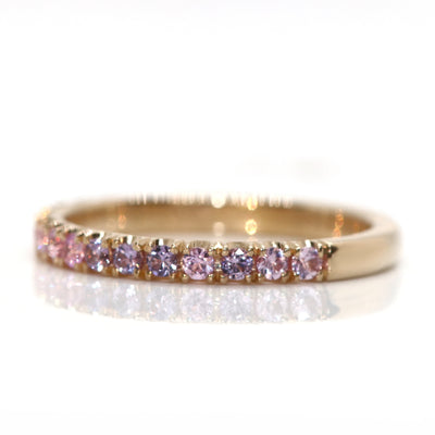 A delicate 14kt gold ring adorned with  brilliant cut pink sapphires in a variety of tones, reflecting beautifully against a white background, shown from the side.