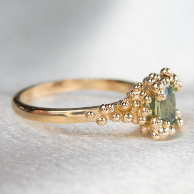 A side view gold ring featuring an oval shaped green sapphire set in an intricate design with gold granules, on a soft, textured surface.