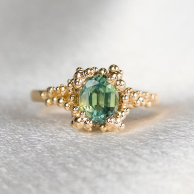 A gold ring featuring an oval shaped green sapphire set in an intricate design with gold granules, on a soft, textured surface