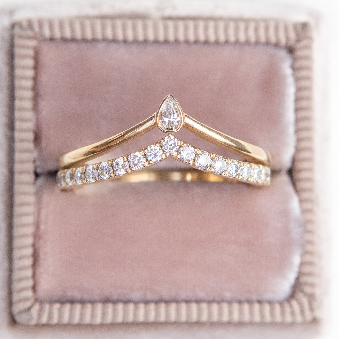 A unique 14kt gold ring featuring a double peak design, adorned with sparkling pave diamonds and a pear diamond centerpiece stacked with a large oval lab diamond solitaire engagement ring shown nestled in a light pink velvet ring box.