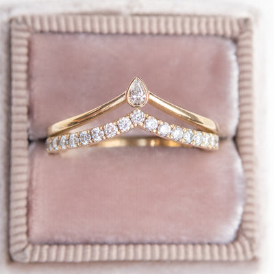 A unique 14kt gold ring featuring a double peak design, adorned with sparkling pave diamonds and a pear diamond centerpiece stacked with a large oval lab diamond solitaire engagement ring shown nestled in a light pink velvet ring box.
