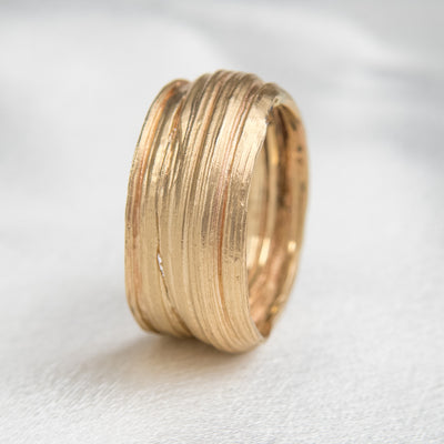 Gold textured ring with layered and organic design, resting on it's side on white silk fabric, showcasing a modern and elegant aesthetic.