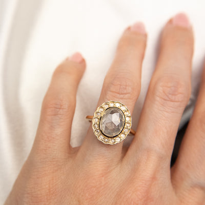 A gold ring with an oval, translucent grey salt and pepper diamond at its center is surrounded by a halo of small round rose cut diamonds, worn on a hand.