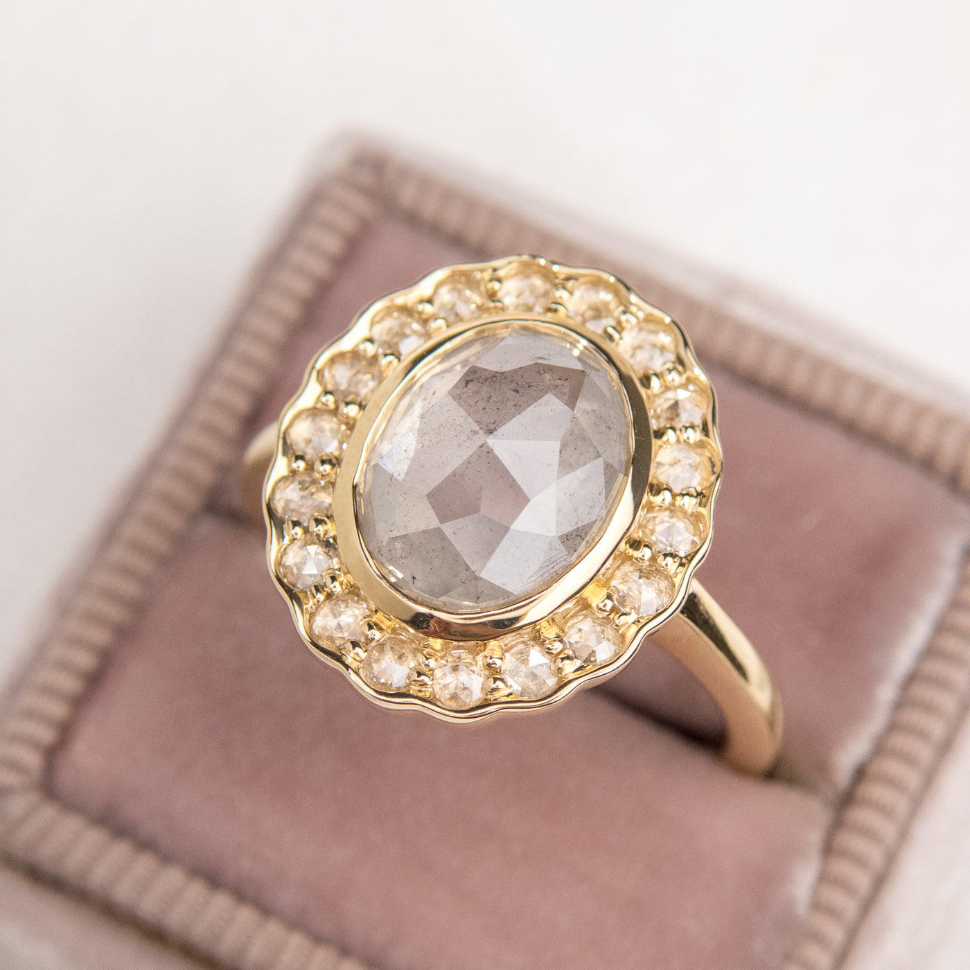 A gold ring with an oval, translucent grey salt and pepper diamond at its center is surrounded by a halo of small round rose cut diamonds, set against a light pink velvet ring box.