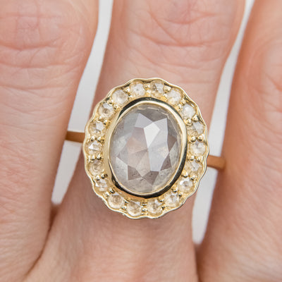 A gold ring with an oval, translucent grey salt and pepper diamond at its center is surrounded by a halo of small round rose cut diamonds, worn on a finger. 