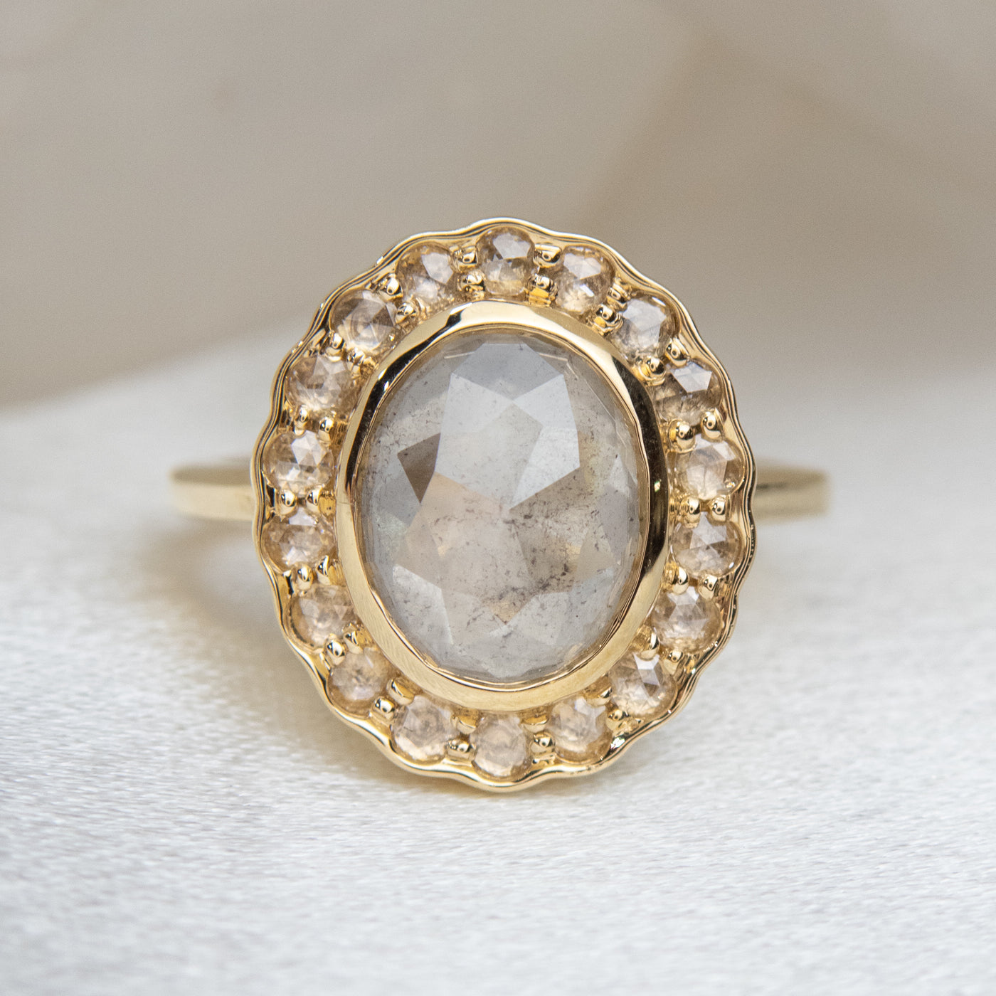 A gold ring with an oval, translucent grey salt and pepper diamond at its center is surrounded by a halo of small round rose cut diamonds, set against a soft, neutral background.