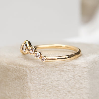 A delicate gold ring with a central pear-shaped diamond, flanked by smaller round diamonds, resting on a soft, textured surface shown from the side.