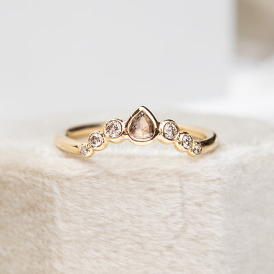 A delicate gold ring with a central pear-shaped diamond, flanked by smaller round diamonds, resting on a soft, textured surface.
