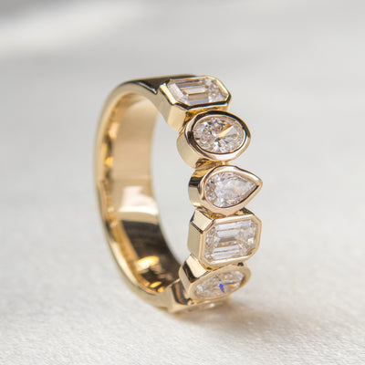 A stylish gold bezel ring adorned with various  lab diamonds, including pear and emerald cuts, resting on it's side on white fabric.