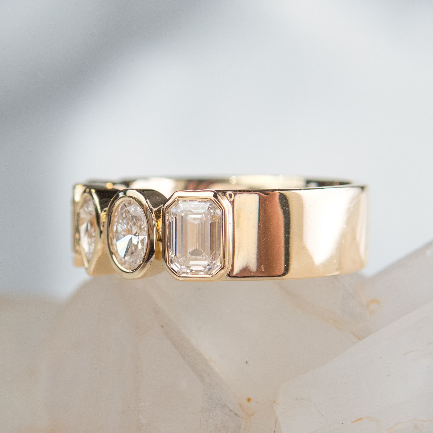 A stylish gold bezel ring adorned with various lab diamonds, including pear and emerald cuts, resting on a crystalline surface shown from the side.