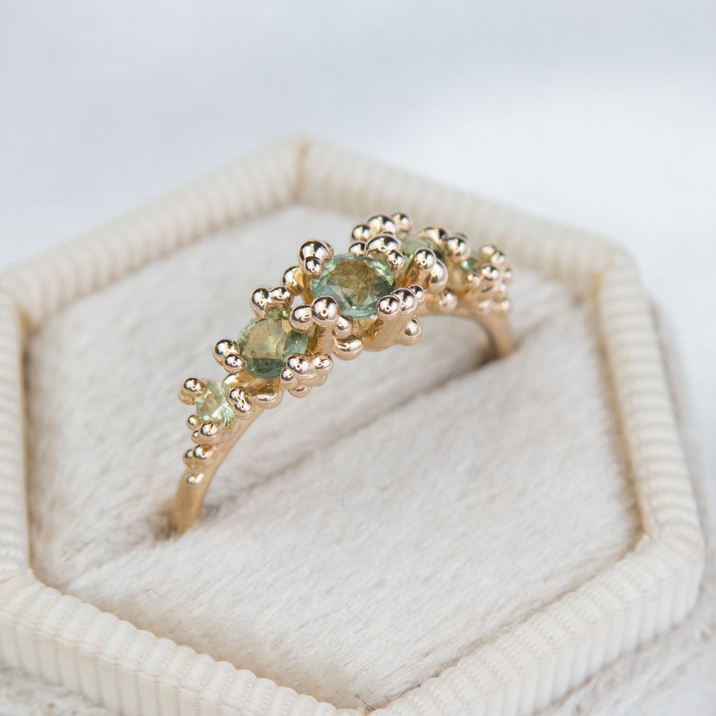 A gold ring adorned with a row five green sapphires set in organic gold granules, in a white fabric ring box. 