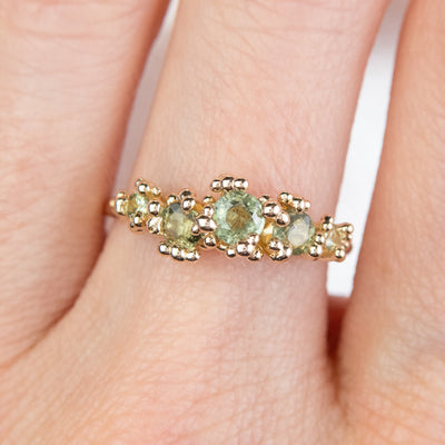 A gold ring adorned with a row five green sapphires set in organic gold granules, worn on a finger.