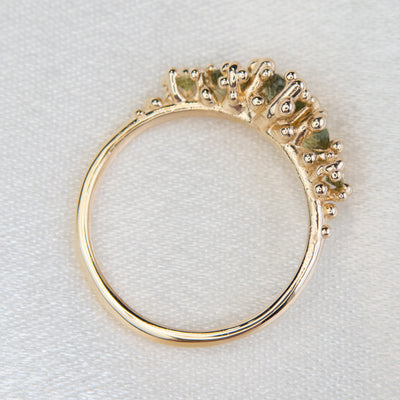 A profile view of a gold ring adorned with a row five green sapphires set in organic gold granules, on a white fabric. 