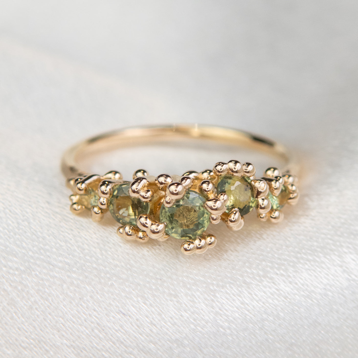 A gold ring adorned with a row five green sapphires, set against a soft white background.