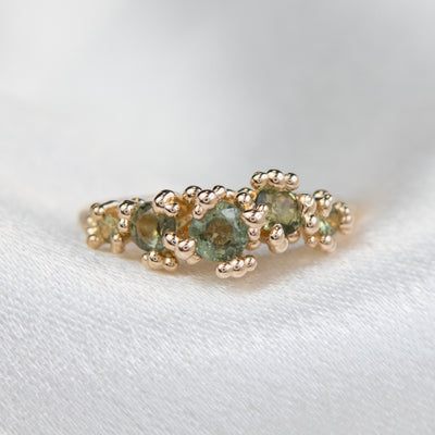 A gold ring adorned with a row five green sapphires, set against a soft white background.