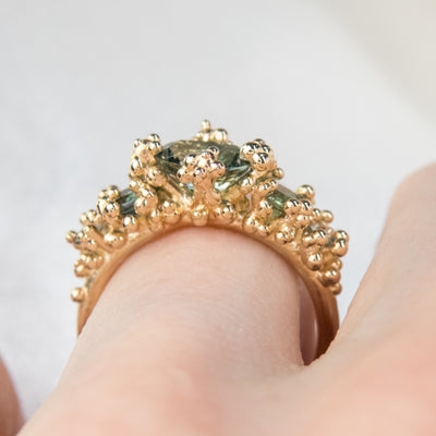 Side profile view of a gold ring featuring five green sapphires, surrounded by textured gold granule accents, worn on a finger.