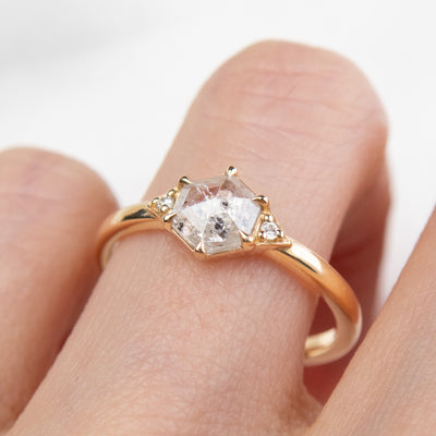 A gold ring with a hexagonal salt and pepper diamond, flanked by small round diamonds on either side, worn on a finger.