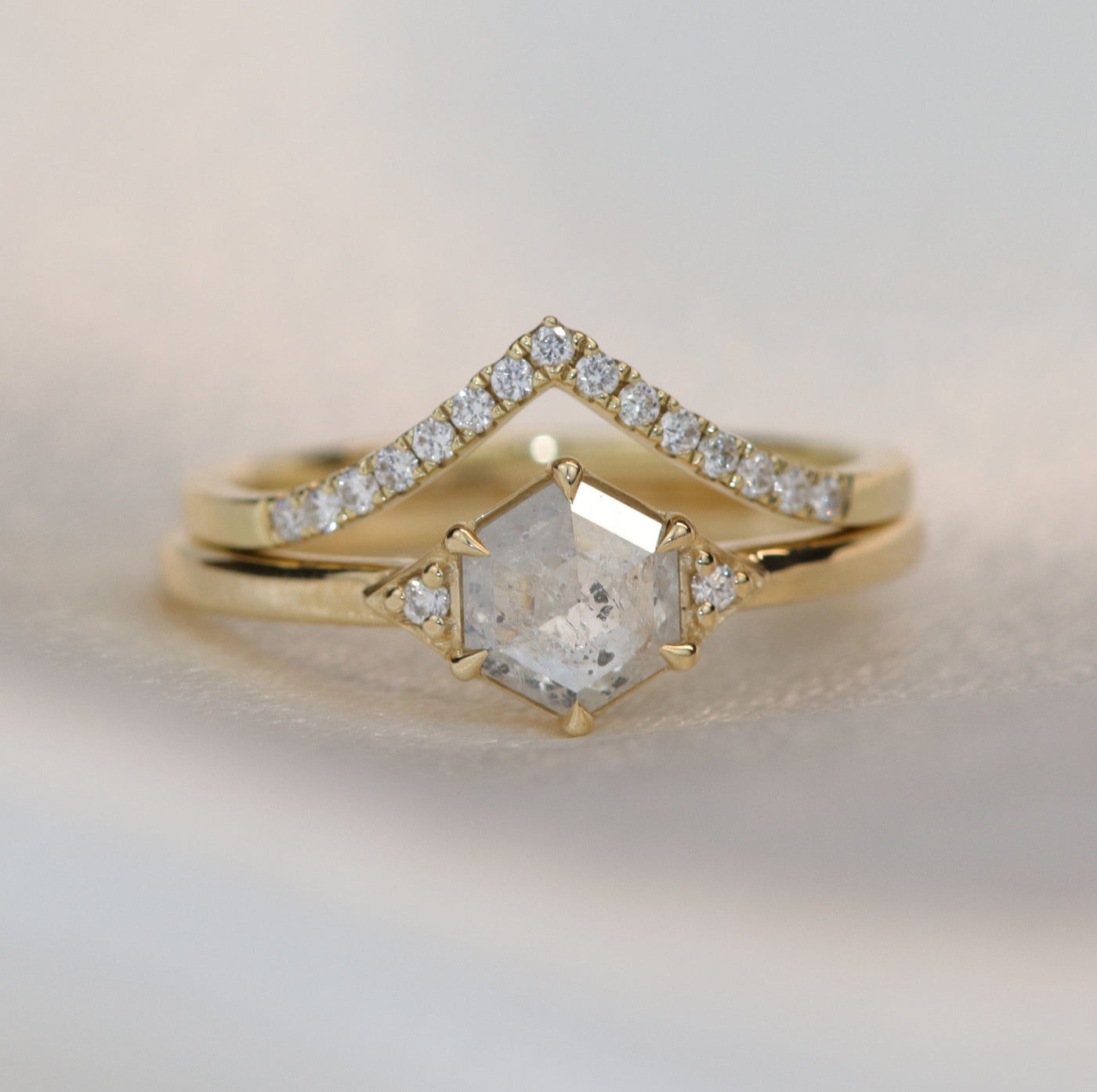 A gold ring with a hexagonal salt and pepper diamond, flanked by small round diamonds on either side, paired with a 'V' shaped diamond band, on a soft white background.
