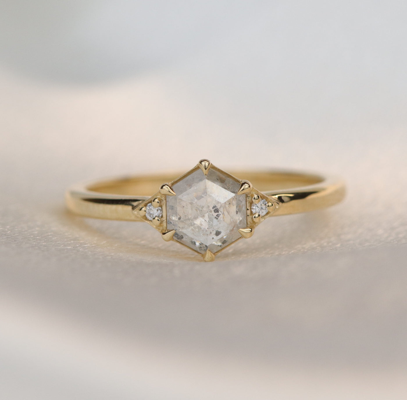 A gold ring with a hexagonal salt and pepper diamond, flanked by small round diamonds on either side, on a soft white background.