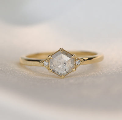 A gold ring with a hexagonal salt and pepper diamond, flanked by small round diamonds on either side, on a soft white background.