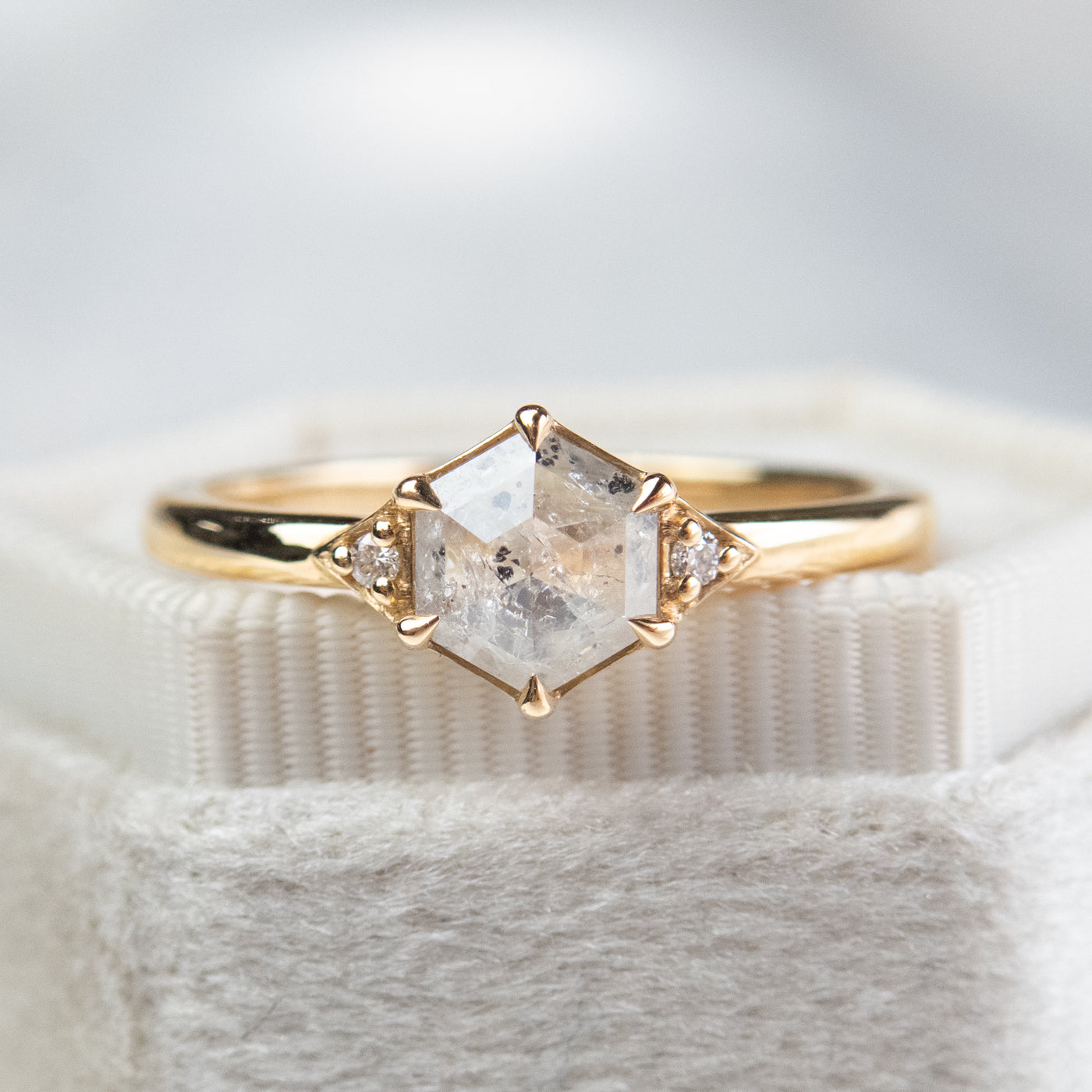A gold ring with a hexagonal salt and pepper diamond, flanked by small round diamonds on either side, rests on a soft, fabric background.