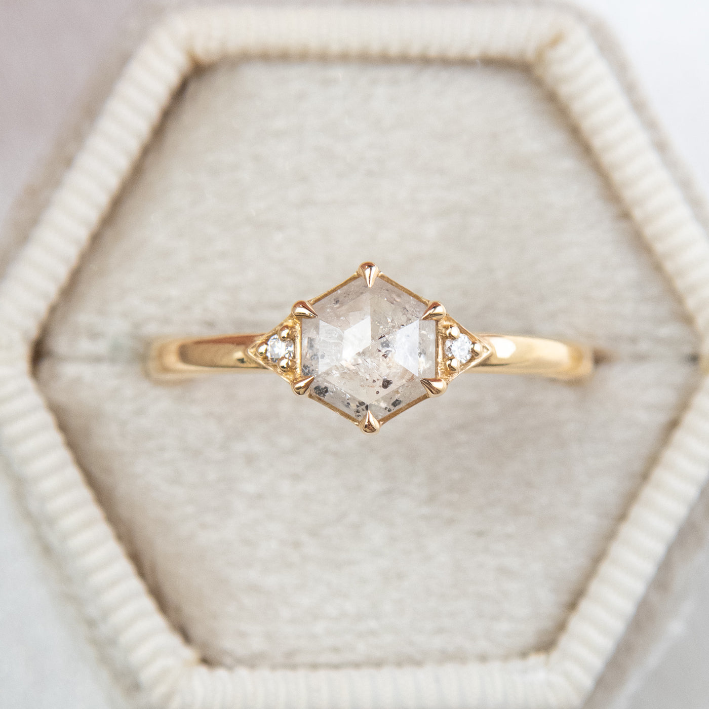 A gold ring with a hexagonal salt and pepper diamond, flanked by small round diamonds on either side, in a hexagonal shaped velvet ring box. 