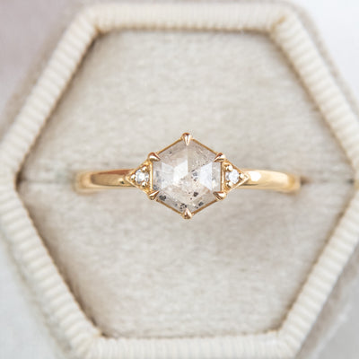 A gold ring with a hexagonal salt and pepper diamond, flanked by small round diamonds on either side, in a hexagonal shaped velvet ring box. 