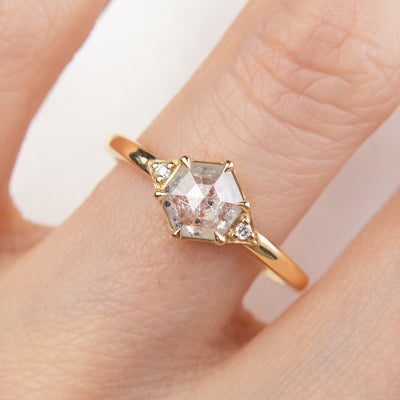 A gold ring with a hexagonal salt and pepper diamond, flanked by small round diamonds on either side, worn on a hand.