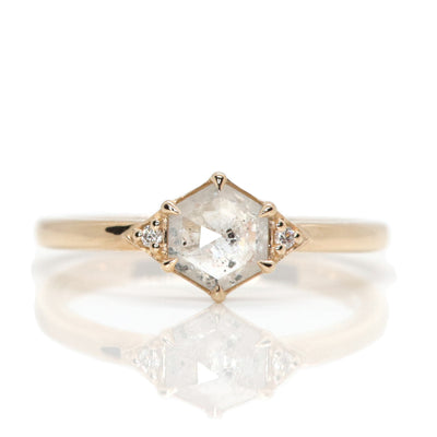 A gold ring with a hexagonal salt and pepper diamond, flanked by small round diamonds on either side, on a reflective white background.
