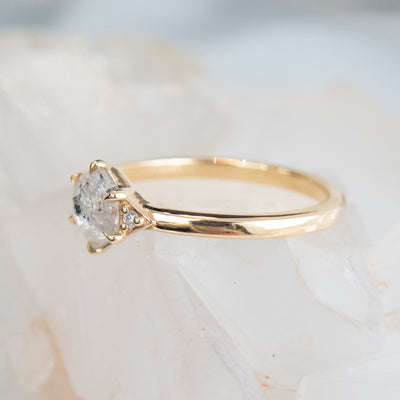 A gold ring with a hexagonal salt and pepper diamond, flanked by small round diamonds on either side, rests on a soft, blurred crystalline background