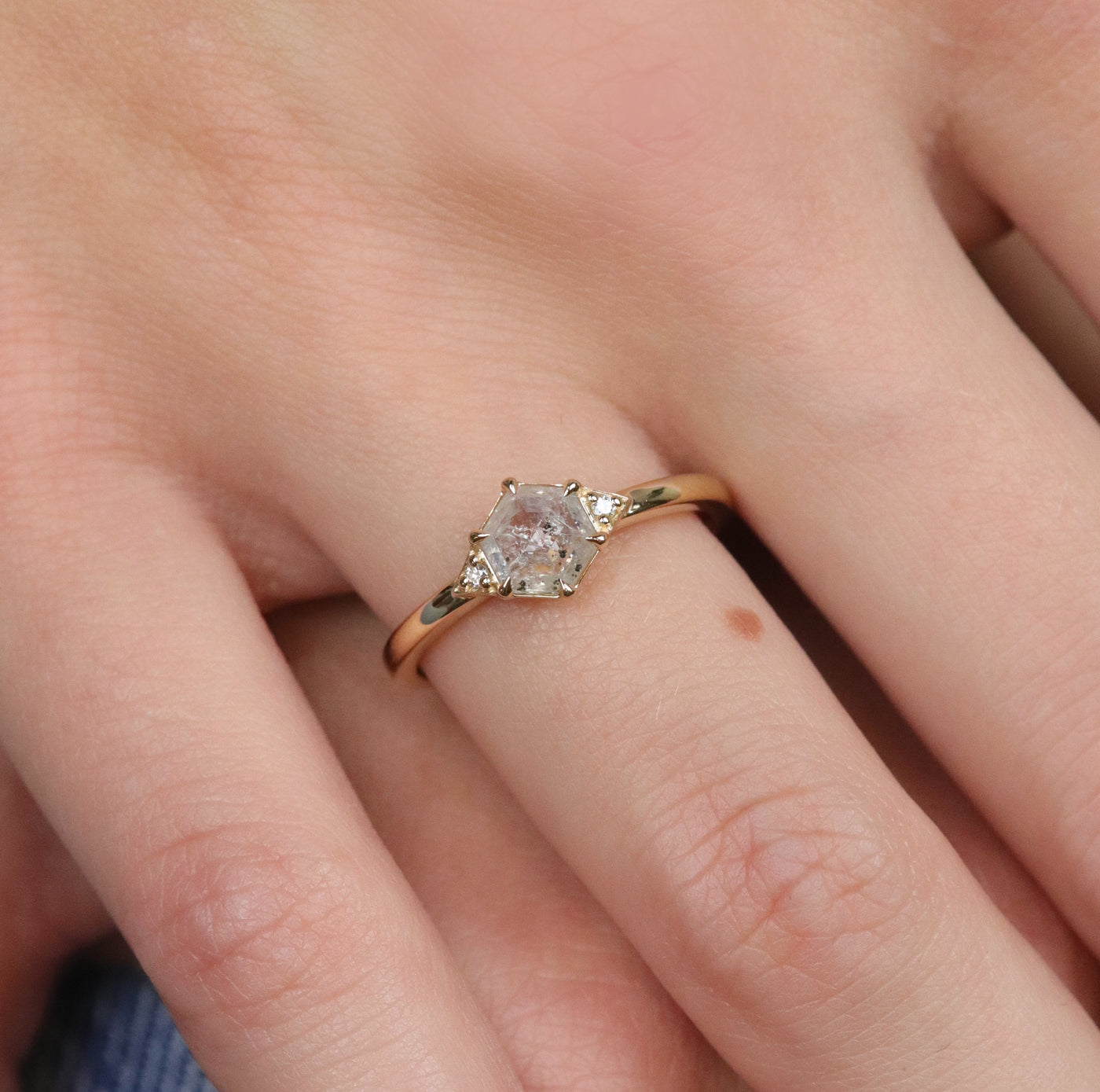 A gold ring with a hexagonal salt and pepper diamond, flanked by small round diamonds on either side, worn on a hand.