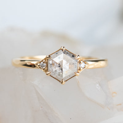 A gold ring with a hexagonal salt and pepper diamond, flanked by small round diamonds on either side, rests on a soft, blurred crystalline background