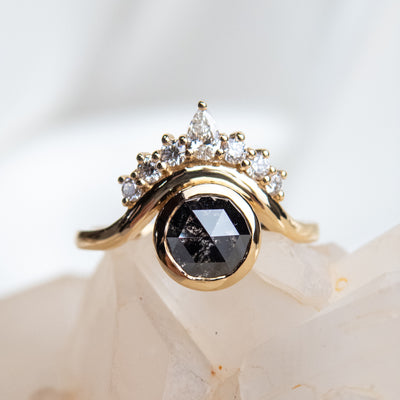 A gold ring with a dark round rose cut salt and pepper diamond, framed by an offset curved band and white diamond accents, set against a light crystalline background. 