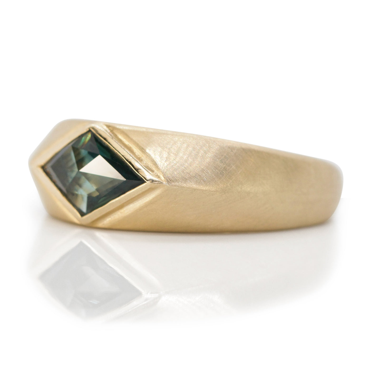 A sleek gold signet ring features a kite, rosecut blue sapphire shown from the side resting elegantly on a white reflective surface.