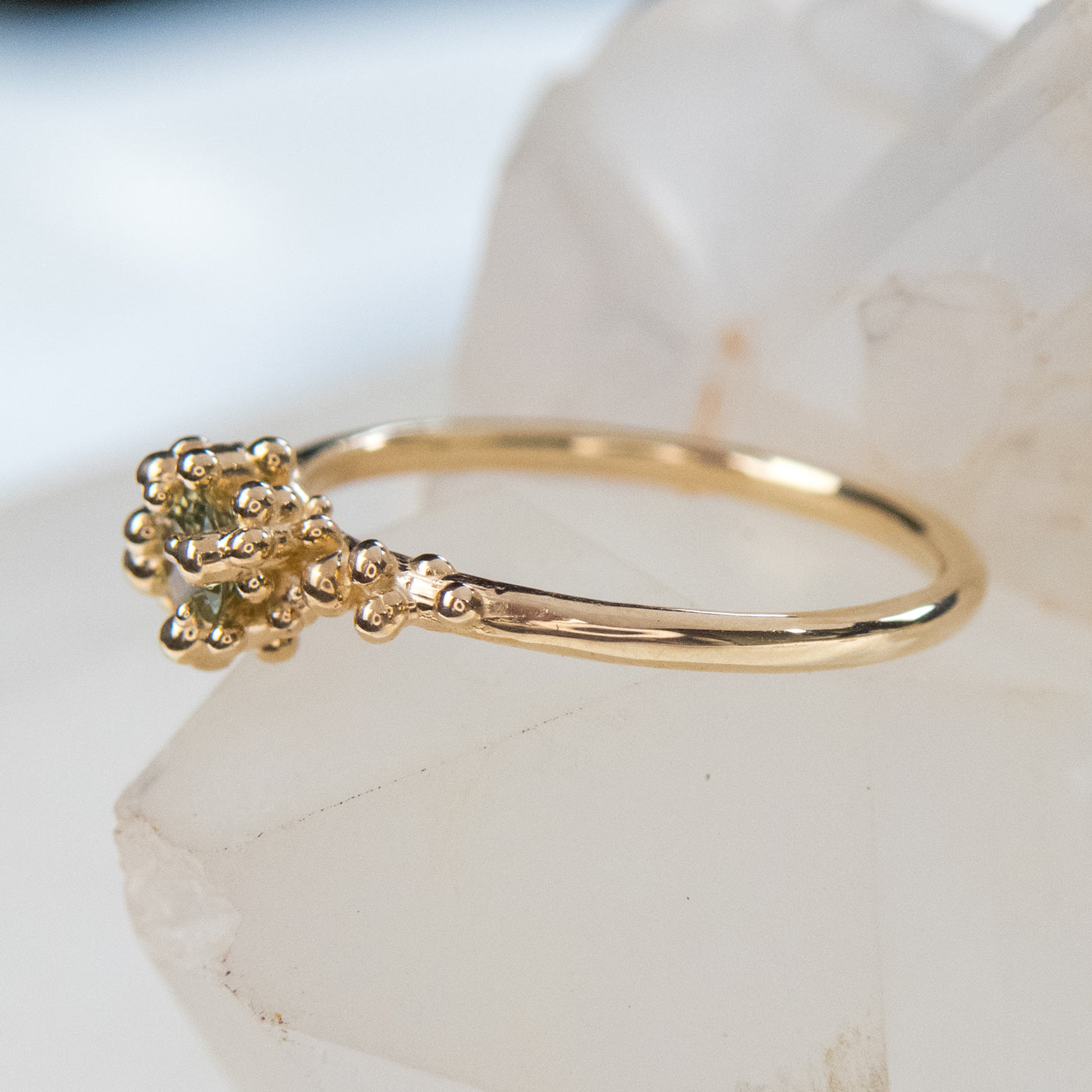 Side view of a gold ring with a cluster of small round gold beads holds a green sapphire at the center, displayed on a white crystalline surface.