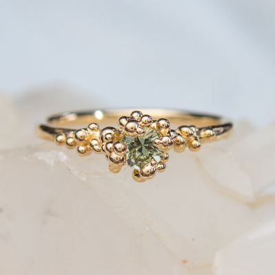 A gold ring with a cluster of small round gold beads holds a green sapphire at the center, displayed on a white crystalline surface.