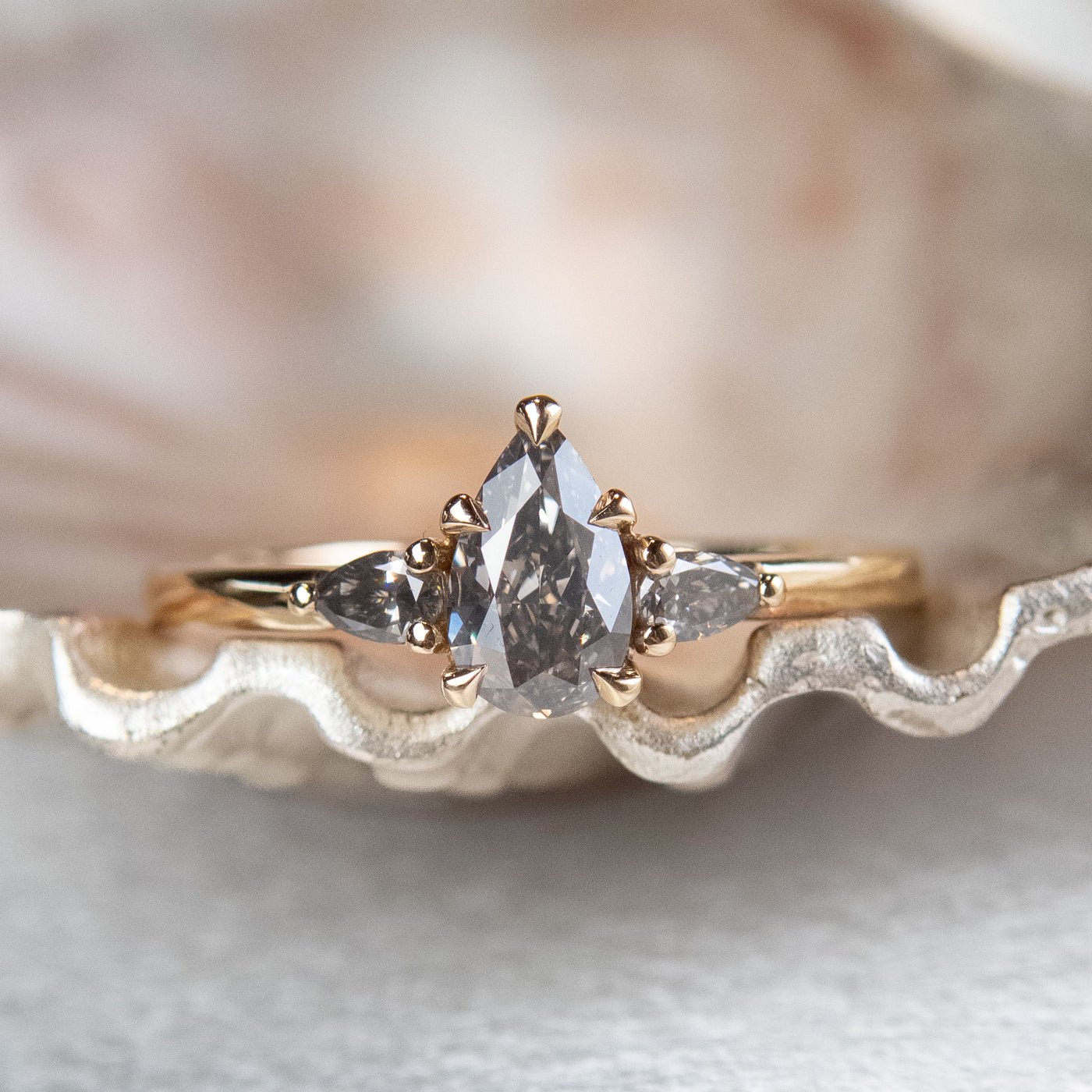 A gold three-stone ring with a fancy grey pear-shaped diamond center and two smaller grey pear-shaped side diamonds on a metallic, textured surface.