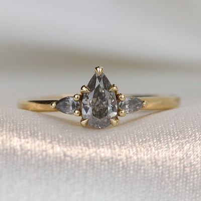 A gold three-stone ring with a fancy grey pear-shaped diamond center and two smaller grey pear-shaped side diamonds on a soft, textured surface.