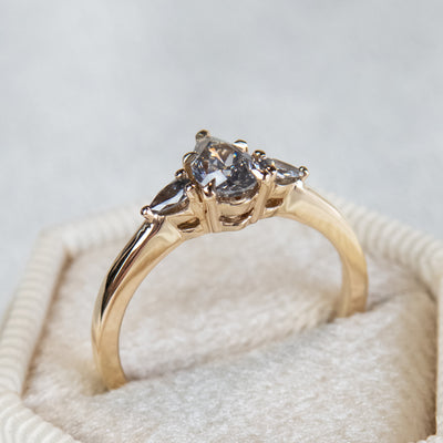 A side profile view of a gold three-stone ring with a fancy grey pear-shaped diamond center and two smaller grey pear-shaped side diamonds on a soft, textured surface.