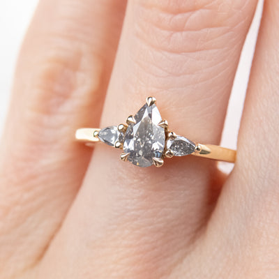 A gold three-stone ring with a fancy grey pear-shaped diamond center and two smaller grey pear-shaped side diamonds, worn on a finger. 