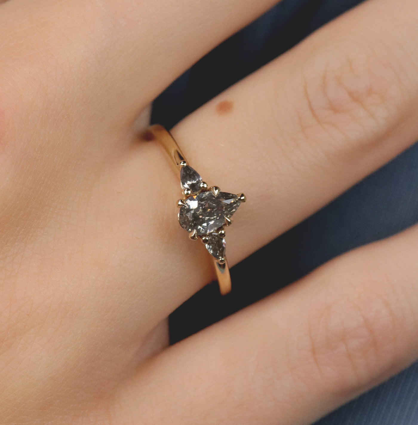 A gold three-stone ring with a fancy grey pear-shaped diamond center and two smaller grey pear-shaped side diamonds, worn on a hand.