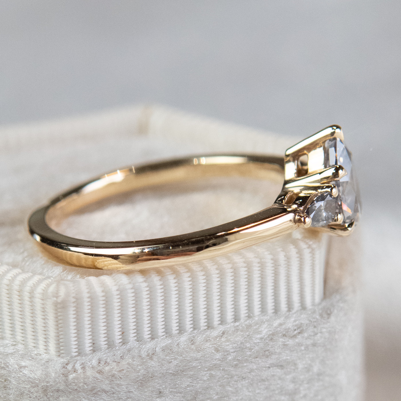 A side profile view of a gold three-stone ring with a fancy grey pear-shaped diamond center and two smaller grey pear-shaped side diamonds on a soft, textured surface.